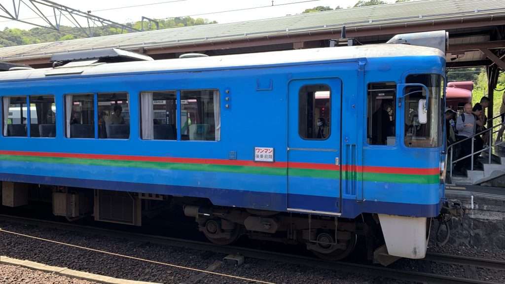 京都丹後鉄道の青い車両を天橋立駅構内で撮影。青い車体に緑と赤のラインが入ったややレトロな列車。運転室横には「ワンマン後乗前降」の案内表示が見える。右側の階段には観光客が並ぶ、晴天の天橋立駅の風景。