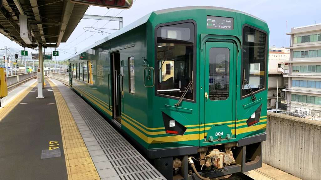 京都丹後鉄道の列車を福知山駅で撮影。福知山から宮津まで乗車した緑色の1両編成の車両。黄色いラインが入った新しいデザインの列車が停車し、左側には駅ホーム、奥には続く線路と市街地が見える曇り空の鉄道風景。