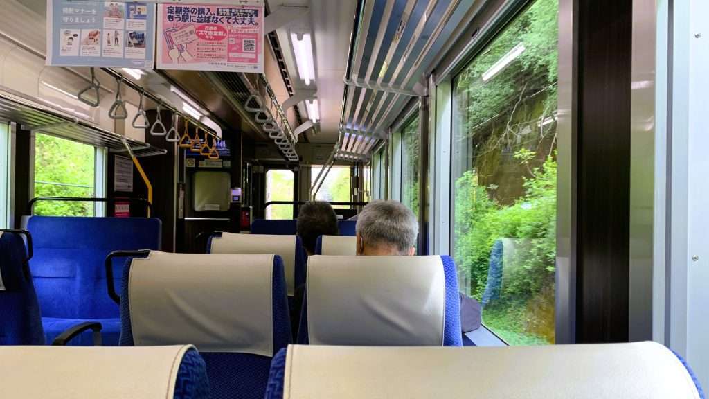京都丹後鉄道の列車内を撮影。福知山から宮津まで乗車した車両の車内風景。青い座席に白いヘッドカバーが付き、つり革や荷物棚が整備された清潔な車内。前方に2人の乗客が見え、窓からは緑豊かな沿線風景が広がる。