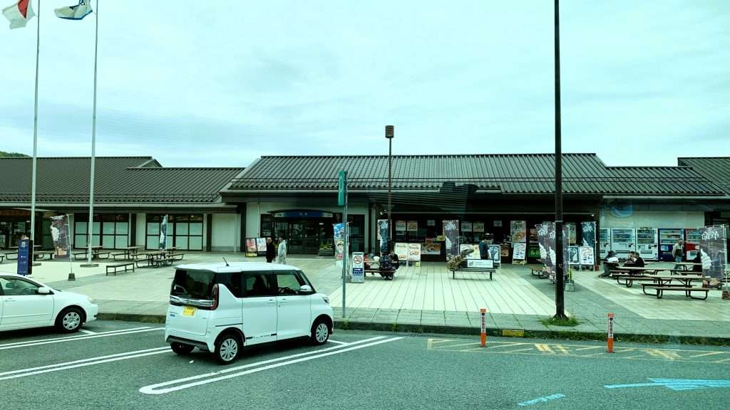 舞鶴若狭自動車道・西紀サービスエリアを撮影。黒い瓦屋根が印象的な低層の建物に店舗や自動販売機が並び、歩道やテーブル、イスのある休憩スペースと駐車場が広がる、曇り空の穏やかなサービスエリア。
