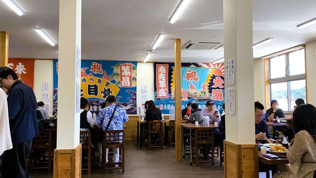 静岡県下田市の市場の食堂 金目亭の店内を撮影。白い壁には大漁旗が飾られ、店内のテーブルは多くの来店客で埋まり、食事を楽しむ人々で賑わう。床や柱には木材が使われ、窓から自然光が差し込む明るい食堂の風景。