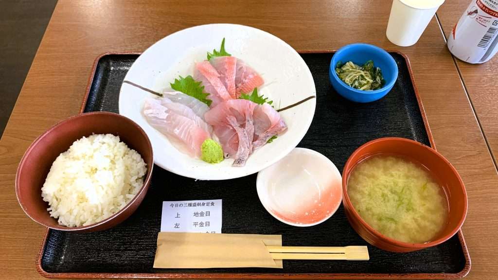 静岡県下田市の「市場の食堂 金目亭」の今日の三種盛刺身定食を撮影。地金目・平金目・はし金目の刺身3種を盛り合わせた丸皿を中心に、ご飯、みそ汁、小鉢、箸、小皿が黒いプレートに並ぶ下田名物の海鮮料理。