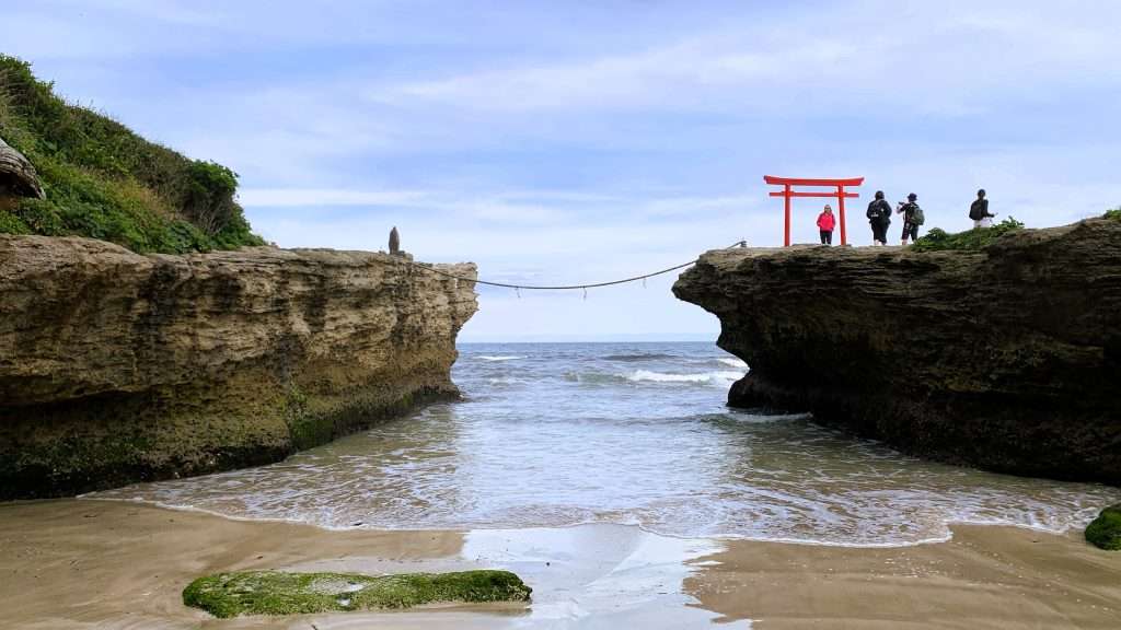 静岡県下田市・白浜海岸にある伊古奈比咩命神社(白濱神社)と歌碑が立つ岩の間の海を撮影。岩の間に海が入り込み、右側には朱色の鳥居と記念撮影をする人々が見える。神社の岩と歌碑の岩は注連縄(しめ縄)で結ばれ、青空と薄い雲が広がる神秘的な海辺の風景。
