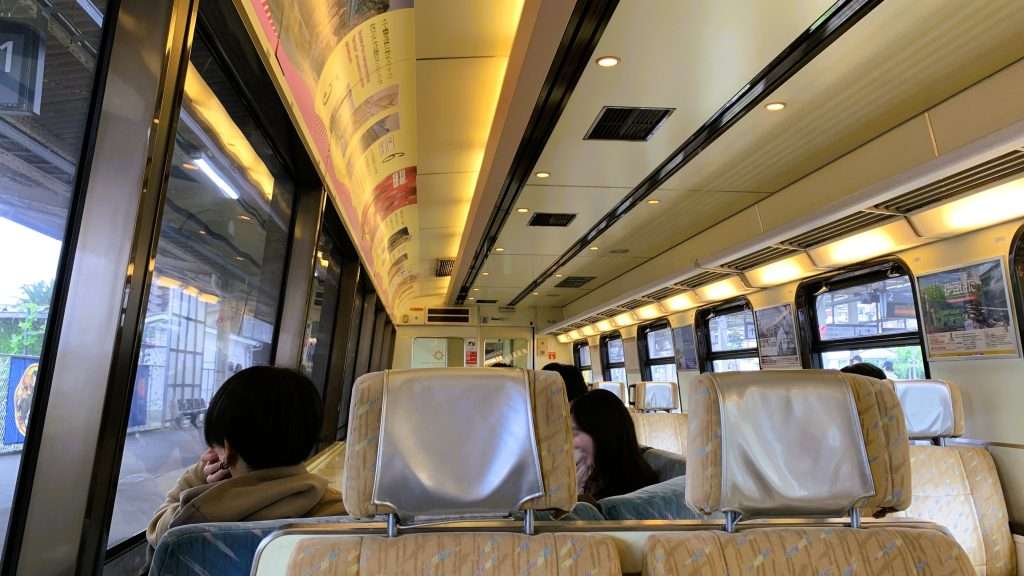 伊豆急行線の黒船電車の車内風景、クリーム色の座席に銀色のヘッドカバーが付いた展望しやすい座席配置。前方には数人の乗客が見え、車窓から駅や沿線風景を楽しめる明るい列車内。