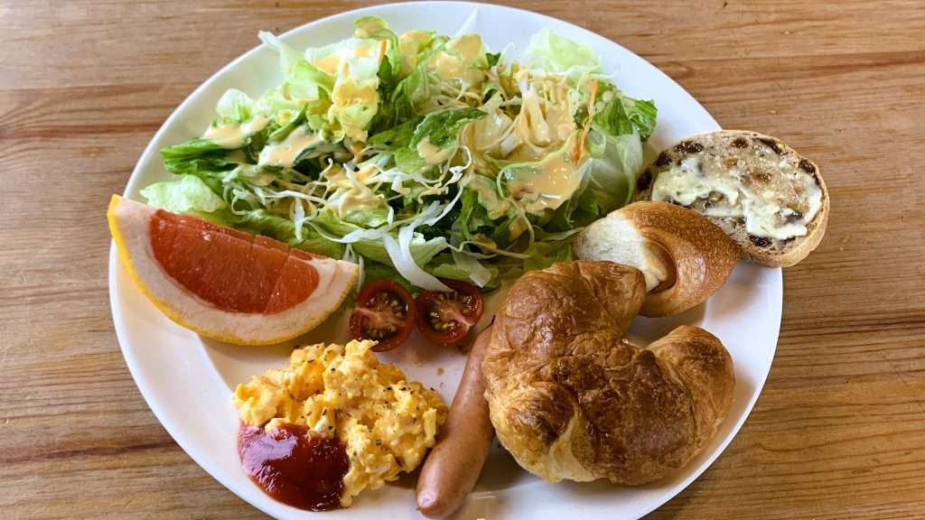 静岡県下田市・白浜海岸近くのジャスミンロッジで提供された朝食を撮影。白い丸皿にサラダ、パン、ソーセージ、卵料理、クロワッサン、グレープフルーツ、ミニトマトが彩りよく盛られた洋食モーニング。木製テーブルに並ぶ温かみのある朝食風景。