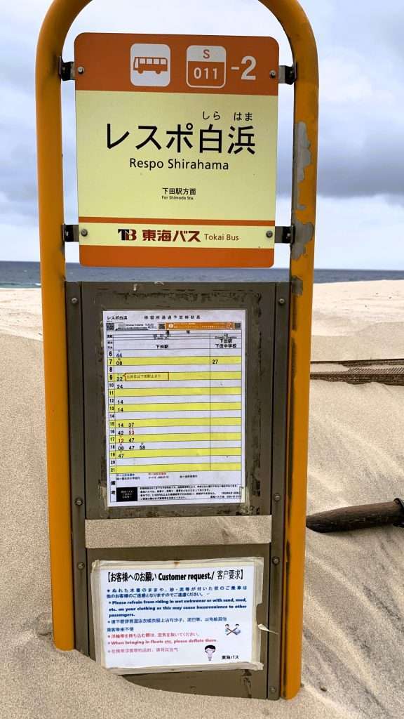 静岡県下田市・白浜海岸近くの砂に埋まったバス停を撮影。「レスポ白浜」と書かれたオレンジ色のバス停標識の下部が白ベージュ色の砂に覆われ、中央には時刻表が掲示されている。奥には白浜海岸の砂浜と海が見える曇り空の風景。
