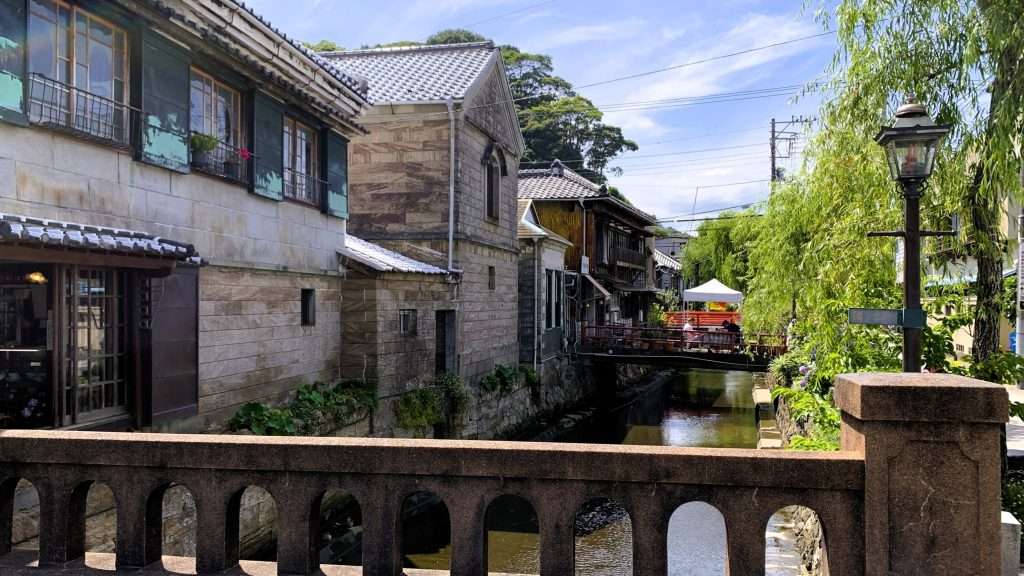 静岡県下田市のペリーロードを撮影。川沿いに石壁の歴史的建造物が並ぶレトロな街並みが広がり、中央には穏やかな川、右側には柳並木が続く。晴れた日に散策を楽しめる下田の歴史的な風景。