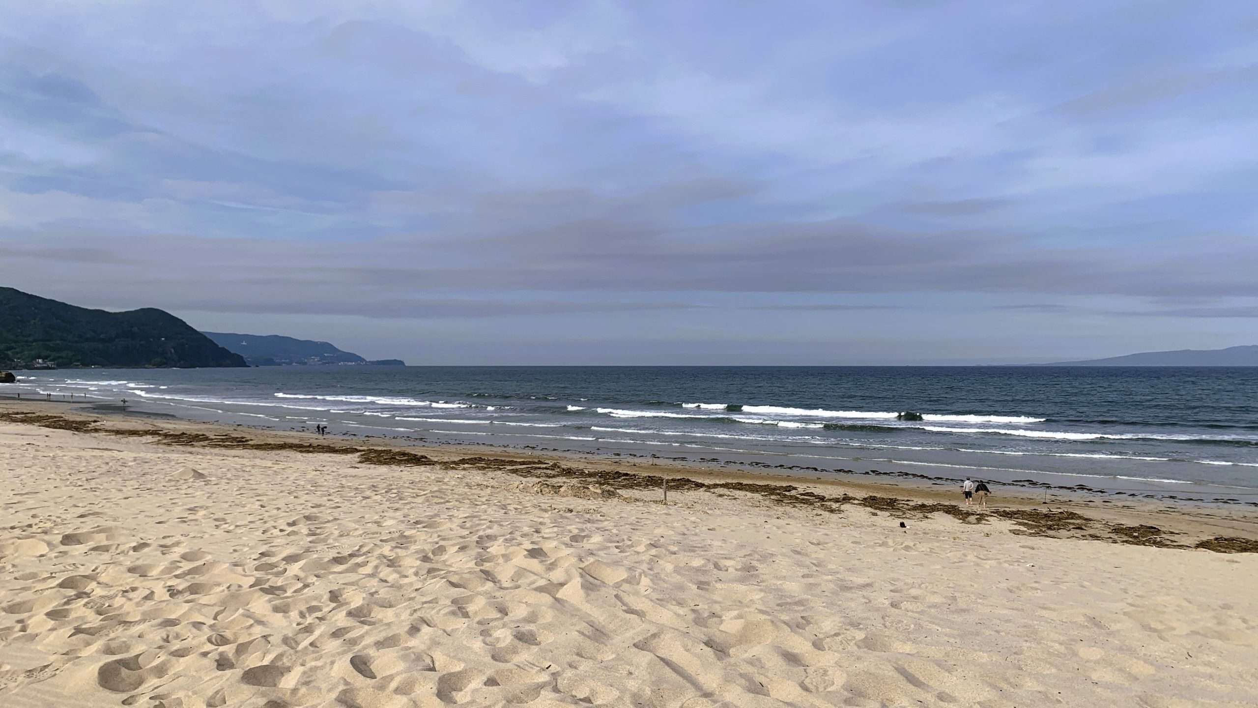 静岡県下田市・白浜海岸を午後に撮影した風景。手前には白い砂浜が広がり、奥には青い海と波が見える。海辺を歩く人の姿が点在し、左側には山が広がる。薄い筋状の雲が浮かぶ空の下、伊豆の美しい海岸景観。