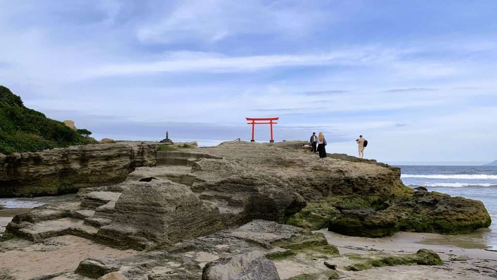 下田市白浜海岸の名所・伊古奈比咩命神社(白濱神社)の海上鳥居を撮影。大岩の上に立つ赤い鳥居と青い海、波の風景が美しく調和し、鳥居前では観光客が記念撮影をしている、静岡県伊豆半島の絶景スポット。