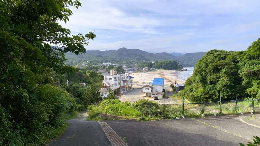 下田市白浜海岸を望む展望風景。山道を出た先から見える白い砂浜と青い海、周辺の建物、背景の山々が調和した、晴れた日の静岡県伊豆半島の自然豊かな景色。